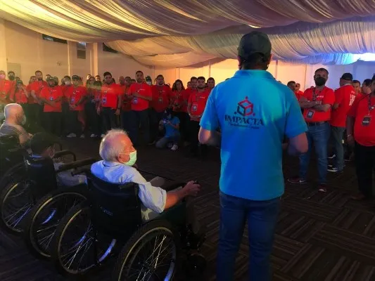 Coach de camiseta azul parado junto a un adulto mayor que se encuentra sentado en una silla de ruedas junto a otros frente a un grupo de personas.