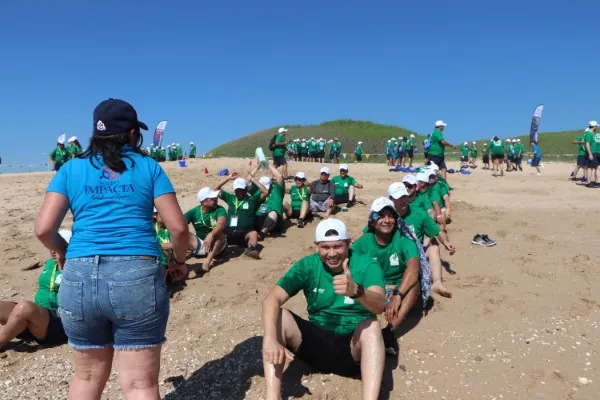 Grupo de personas con playeras verdes integrantes de un equipo están sentadas en una fila bajo el sol en la arena.