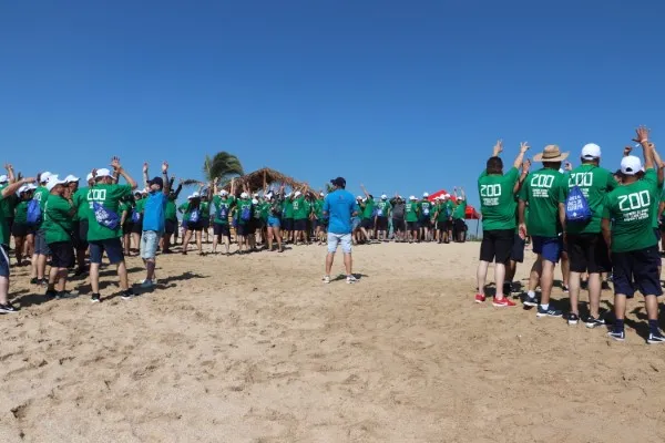 Grupo de Personas reunidas con playeras verdes con coaches de playera azul en una integración de equipos en la playa