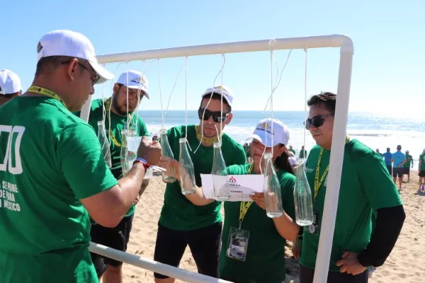 Personas con playeras verdes y gorra son Integrantes de un equipo realizando una dinámica de integración y creatividad con botellas de plástico en una playa.