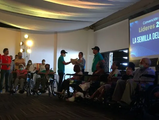 Personas en un evento donde hay adultos mayores sentados en sillas de ruedas y alguien entrega un reconocimiento durante la ceremonia.